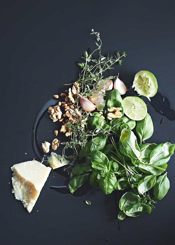 Kräuter, Knoblauch, Parmesan und weitere Zutaten für grünes Pesto auf schwarzem Untergrund - Foodfotografie